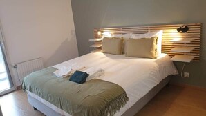 A neatly made bed with a green throw blanket and pillows, a bedside table with a lamp, and a wall-mounted shelf with a hat.