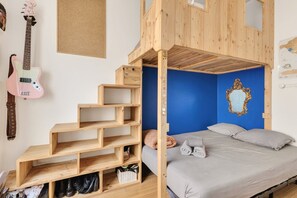 Uma cama beliche com escada de madeira, uma guitarra rosa e uma parede azul com espelho decorativo.