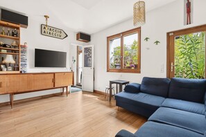 Uma sala de estar com um sofá azul, piso de madeira, uma televisão e uma janela com vista para áreas verdes.