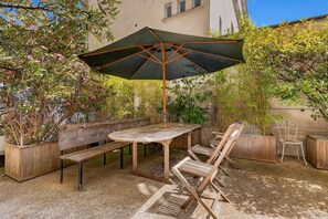 An outdoor patio with a wooden table and chairs, a metal umbrella, and various plants.