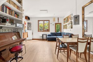 Un vaste espace de vie comprenant un piano, un canapé bleu, une table à manger avec des chaises et une bibliothèque remplie de livres et d’objets décoratifs.