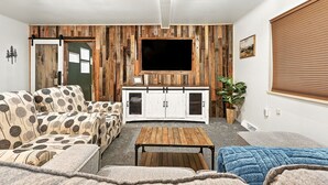 A living room with a wooden wall panel, a TV mounted above a white cabinet, and a sofa with a leaf pattern. - Mountain Serenity Retreat - Larkspur Retreat (Larkspur)