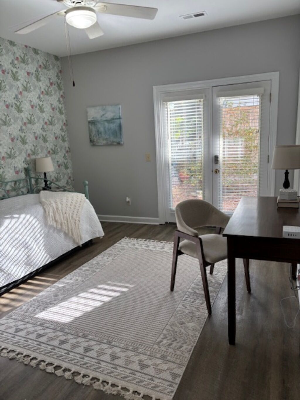 A bedroom with a bed, a chair, a table, a ceiling fan, and a window with blinds.