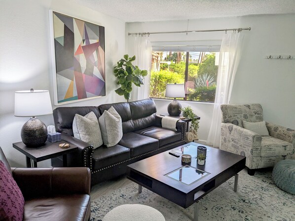 A living room with a leather sofa, a coffee table, a side table with a lamp, and a window with a view of greenery. - Delightful townhome with AC in Palm Springs (Palm Springs)