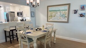 Une salle à manger avec une table en bois dressée pour quatre personnes, entourée de chaises blanches. On aperçoit, au fond, une cuisine avec des armoires blanches et des appareils électroménagers en acier inoxydable.