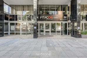 A modern building entrance with glass doors, a sign reading "VITACON 104," and a QR code on the glass.