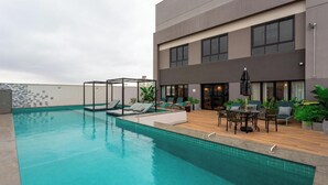 Une piscine sur le toit, dotée de chaises longues, d’un espace salon couvert et d’un bâtiment avec de grandes fenêtres et une porte.