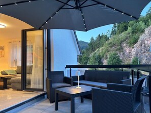 Ein Balkon mit Korbmöbeln, eine Glastür zu einem Wohnbereich und Blick auf einen bewaldeten Hügel.