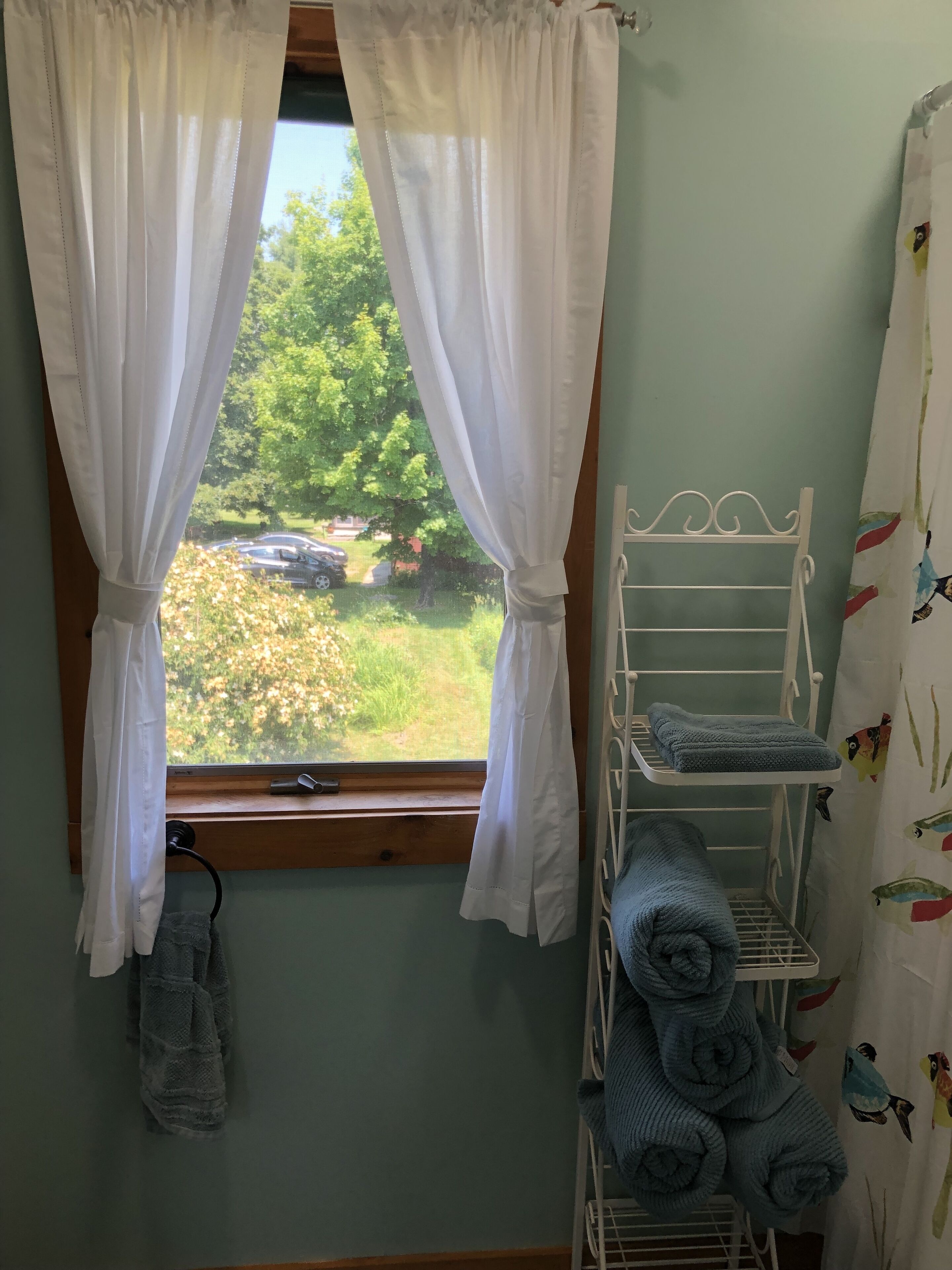 Una habitación con una ventana cubierta por cortinas blancas, un estante metálico blanco con toallas dobladas y una pared con tela estampada de pájaros.