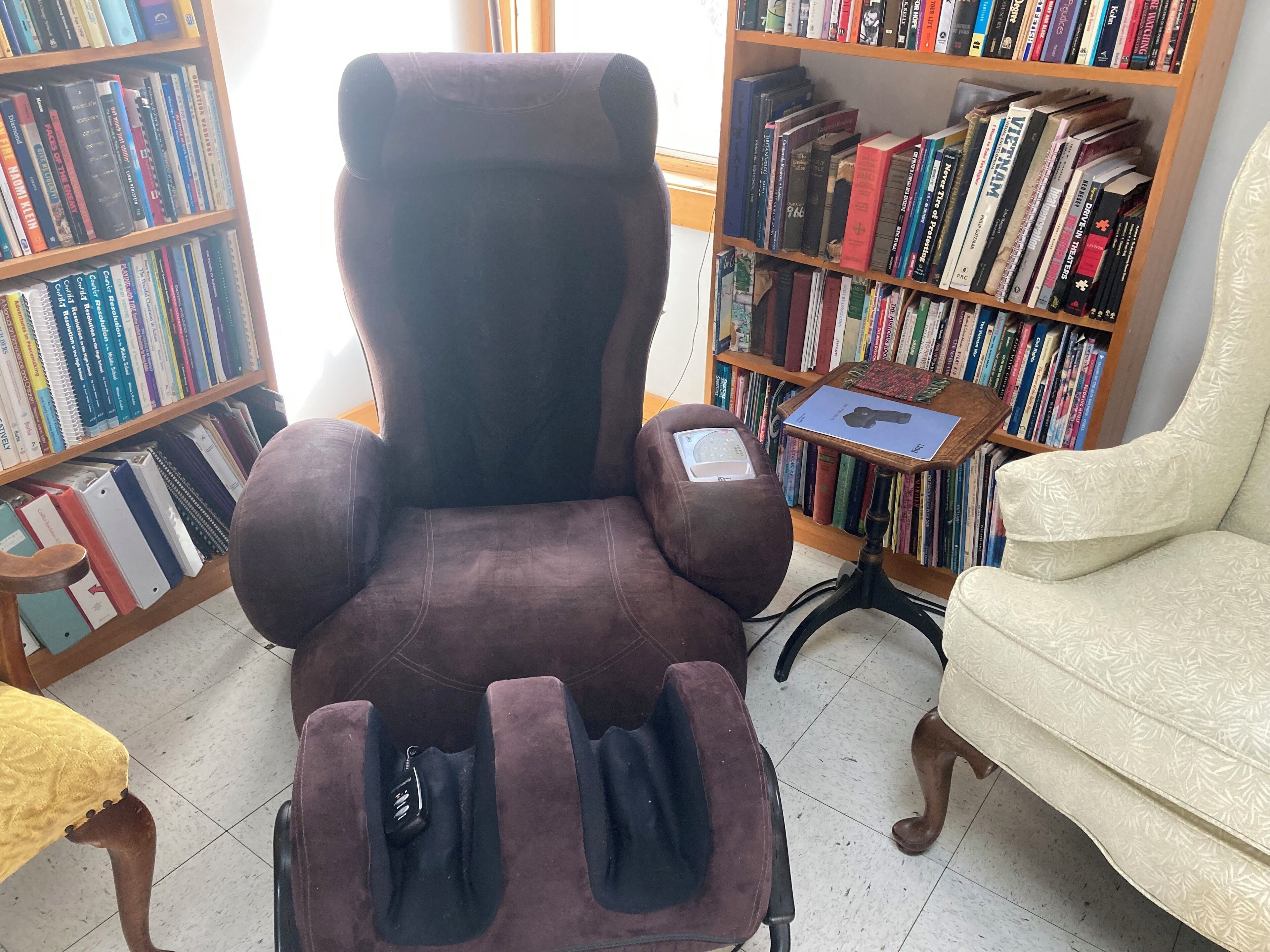 A brown leather chair with a massage pad in a room with bookshelves and a white armchair.
