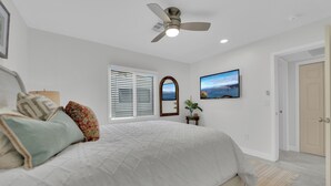Une chambre à coucher avec un lit, un ventilateur de plafond, une fenêtre avec des stores, un miroir encadré et un tableau accroché au mur.