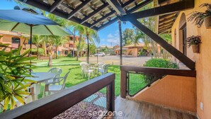 A covered outdoor seating area with tables and chairs, a view of a street, and a building with a tiled roof. - Condominium With Gym and Pool in Guarajuba - Camacari GSH (Camaçari)