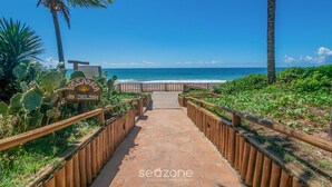 A pathway leading to a beach with a wooden fence, a sign, and palm trees. - Condominium With Gym and Pool in Guarajuba - Camacari GSH (Camaçari)