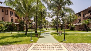A courtyard with palm trees, a pathway, and a brick-paved area leading to a building with balconies. - Condominium With Gym and Pool in Guarajuba - Camacari GSH (Camaçari)