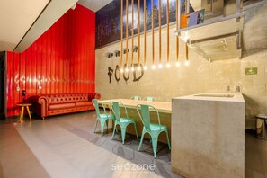 A modern kitchen with a red accent wall, a dining area with a wooden table and teal chairs, and a cityscape mural above the counter.