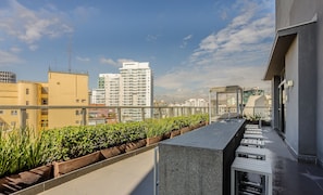Une terrasse sur le toit dotée d’un long espace étroit aménagé avec des sièges, d’un petit élément décoratif aquatique et d’une vue sur les immeubles de grande hauteur environnants.