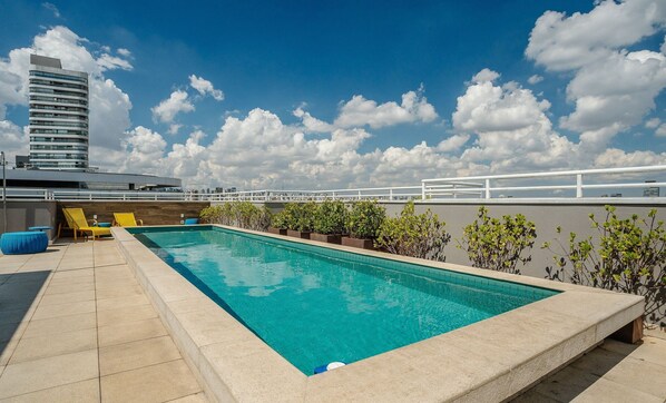 A rooftop pool with a clear blue sky, a tall building in the background, and a white railing.