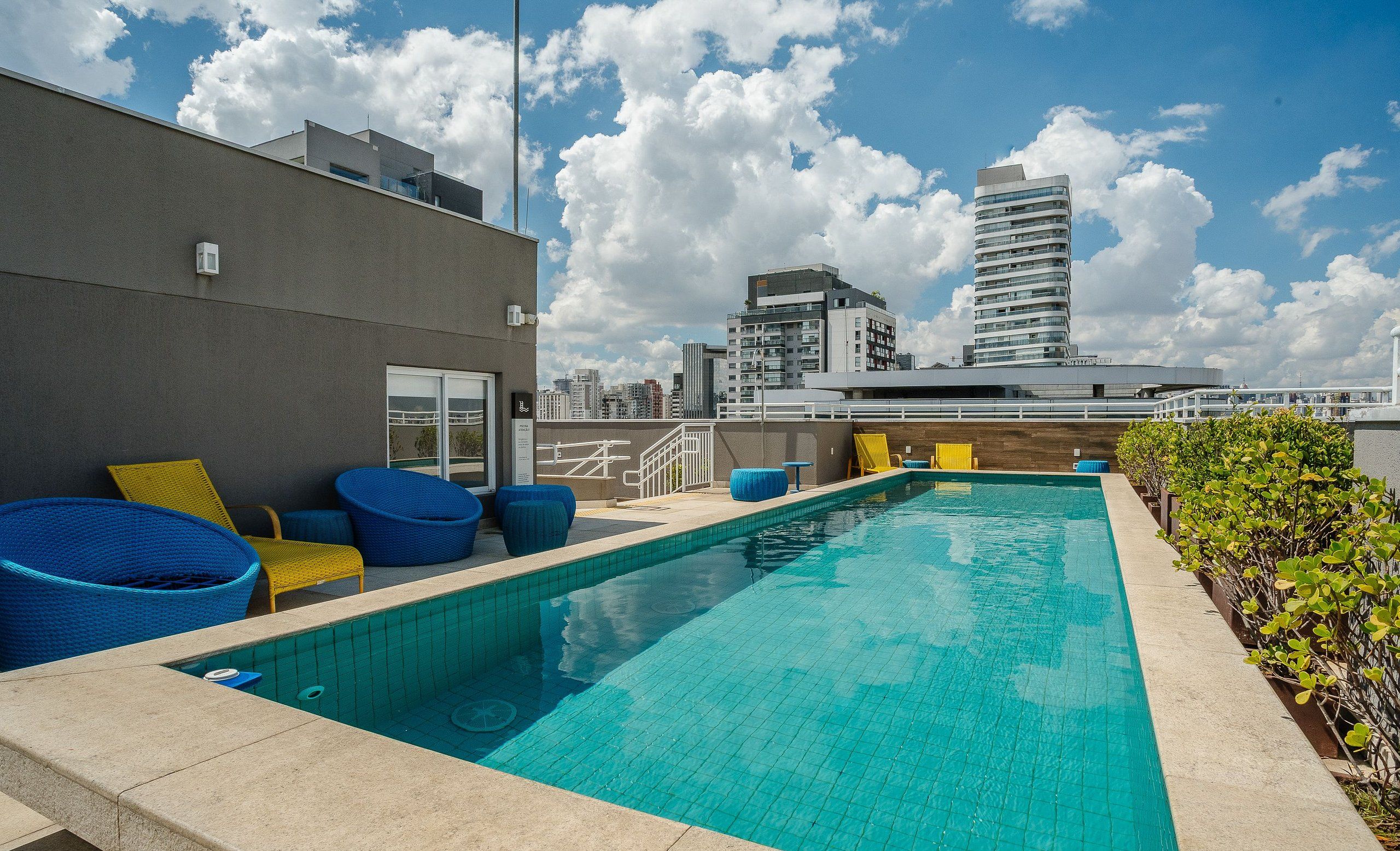 Ein Pool auf dem Dach mit blauen und gelben Liegestühlen, einem klaren blauen Himmel und modernen Gebäuden im Hintergrund.