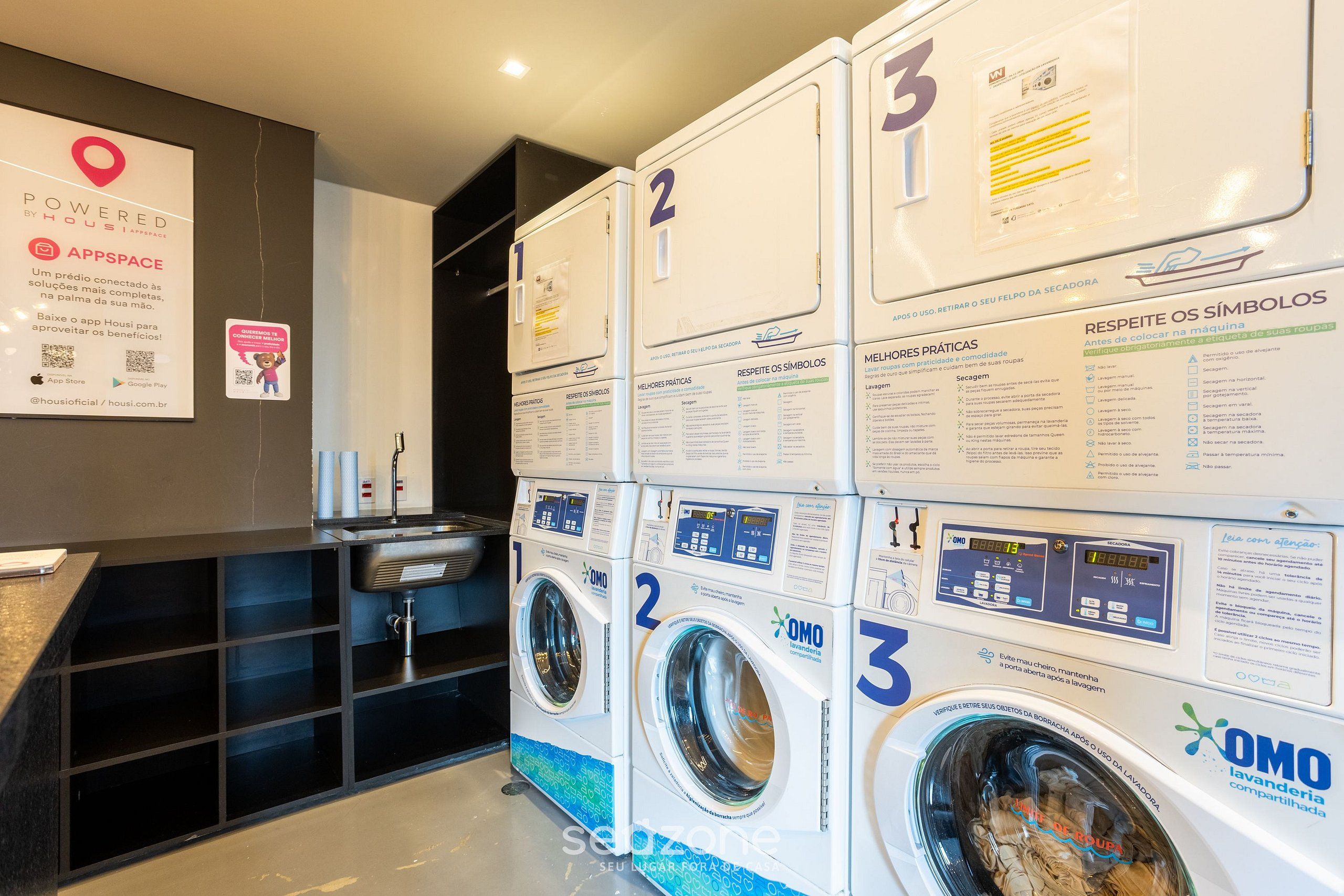 Une buanderie de l’hôtel équipée de trois machines à laver, chacune dotée d’un panneau de commande et d’instructions d’utilisation. Un panneau mural indique l’emplacement grâce à une épingle rouge et fournit des informations sur l’application de l’hôtel.