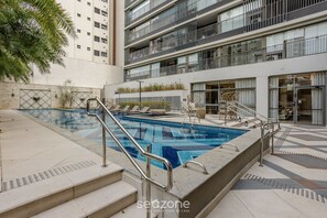 A swimming pool with steps leading to it, surrounded by a building with balconies and a garden area.