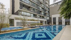 A modern building with multiple balconies, a swimming pool with a geometric tile pattern, and a landscaped area with trees and lounge chairs. - Studio in Condominium With Pool in Paraiso Zah0811 (São Paulo)
