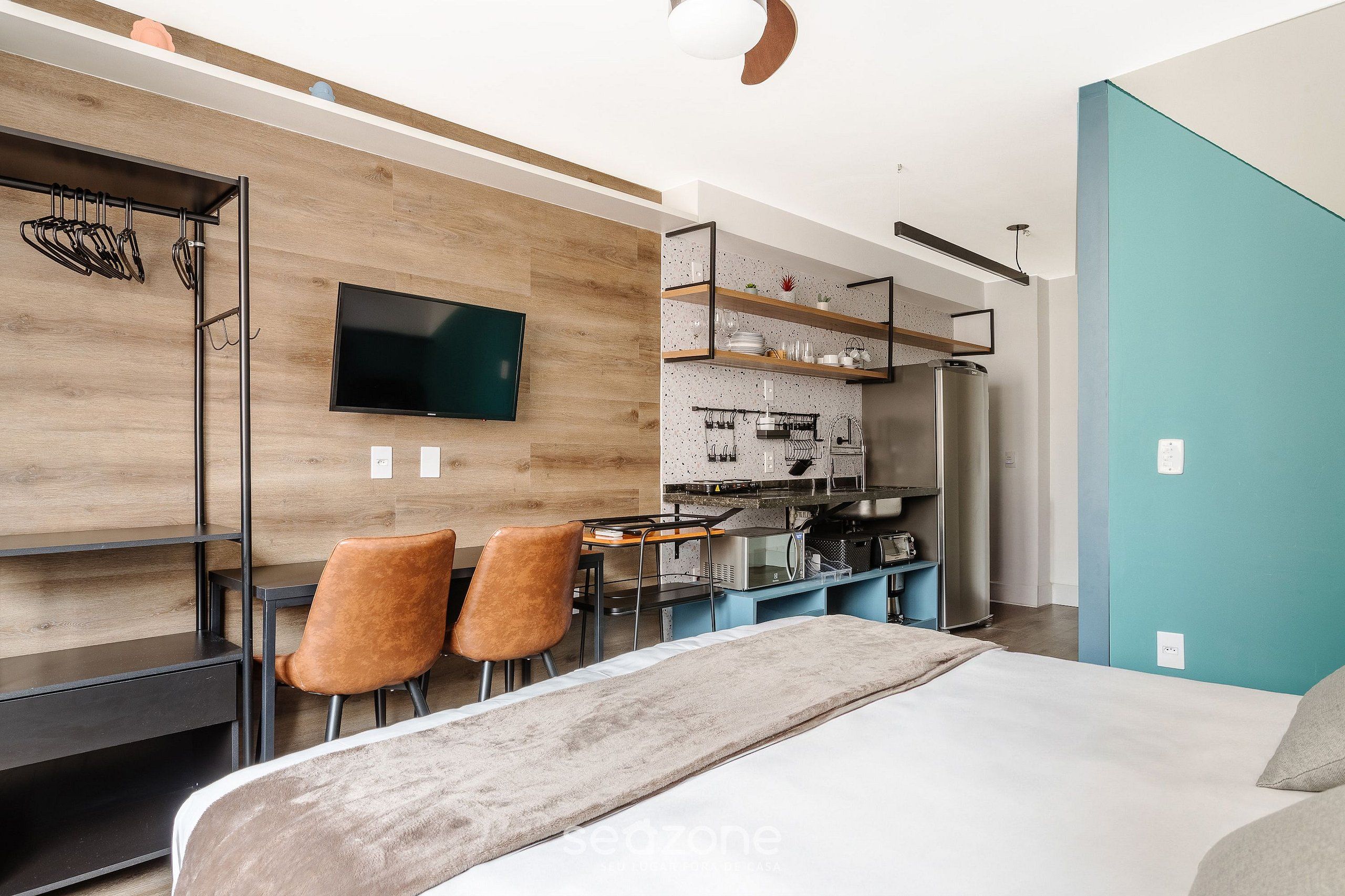 Quarto moderno com uma cama, uma escrivaninha e uma televisão. Há duas cadeiras e um espaço de cozinha com geladeira e micro-ondas.