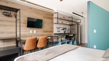 Quarto moderno com uma cama, uma escrivaninha e uma televisão. Há duas cadeiras e um espaço de cozinha com geladeira e micro-ondas.