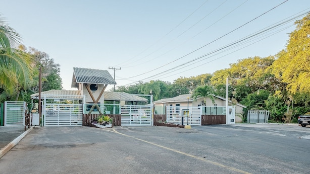 A gated entrance with a unique architectural structure, a parking area, and lush greenery. - Cond. Parque das Arvores - PDA (Camaçari)