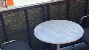 A round table with a white marble top and ornate legs, accompanied by two black chairs, set against a textured wall with a garage door in the background. - WatzmannEG (Augsburg)