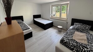 A bedroom with a bed, a small table, a radiator, and a window with a view of greenery. - WatzmannEG (Augsburg)