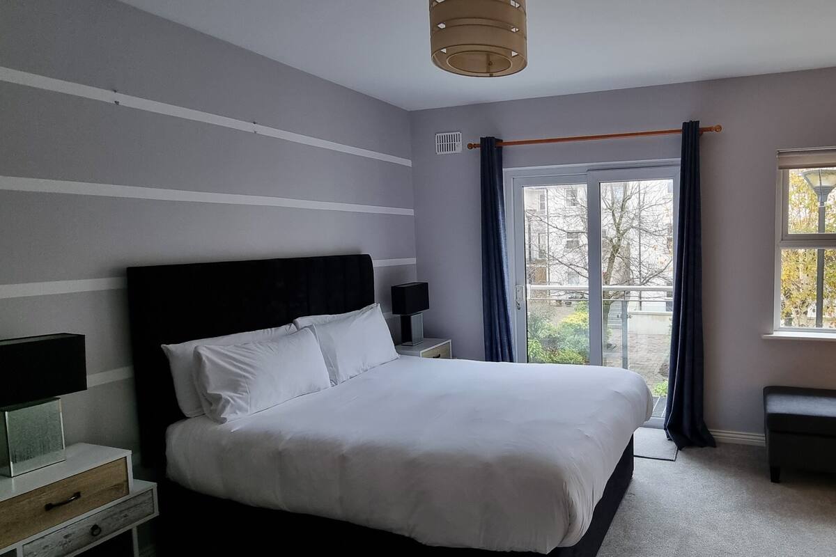 A bedroom with a large bed, a nightstand with a lamp, a dresser, and a window with a view of trees.