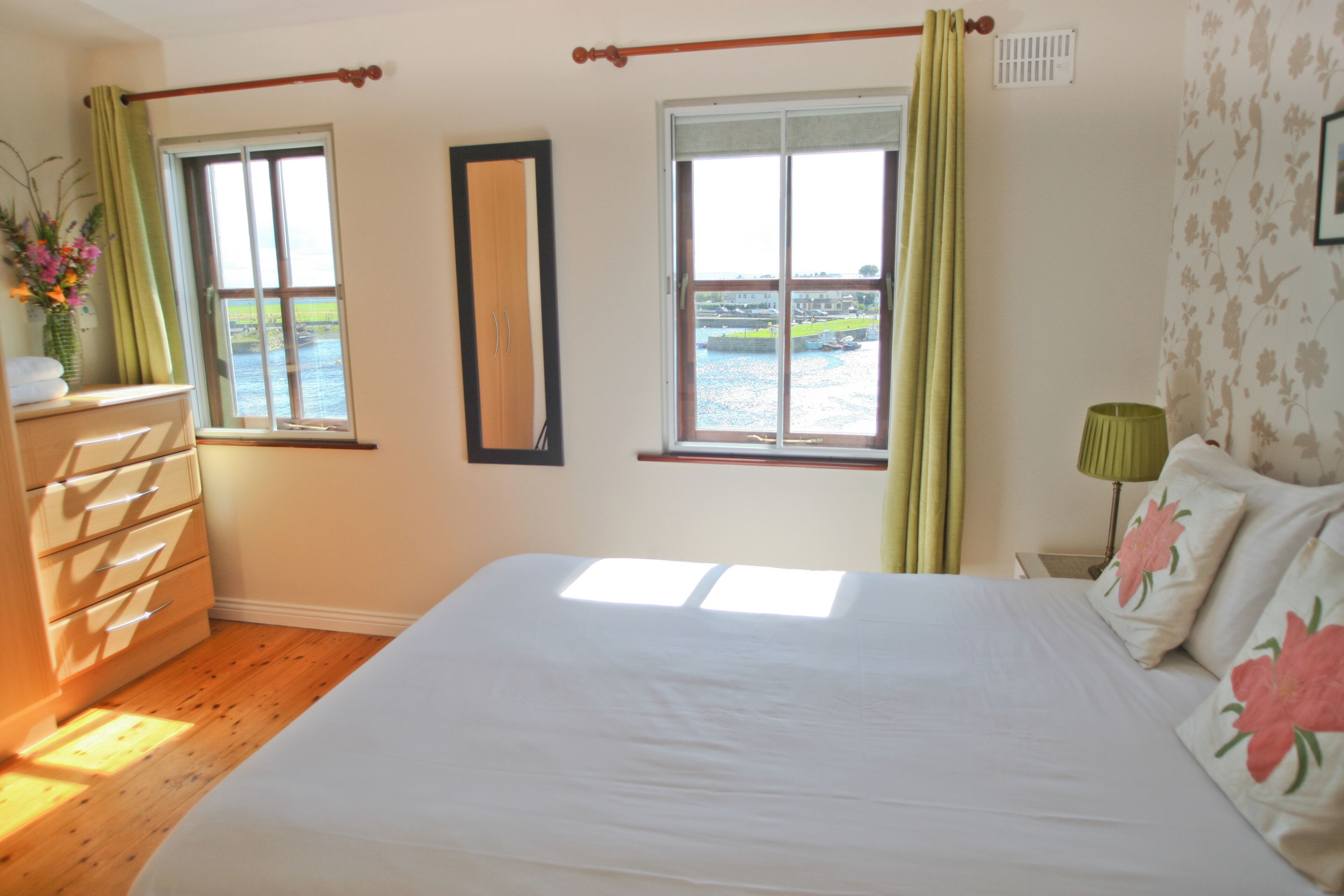 Un dormitorio con una cama grande, dos ventanas con cortinas, un aparador y un espejo.