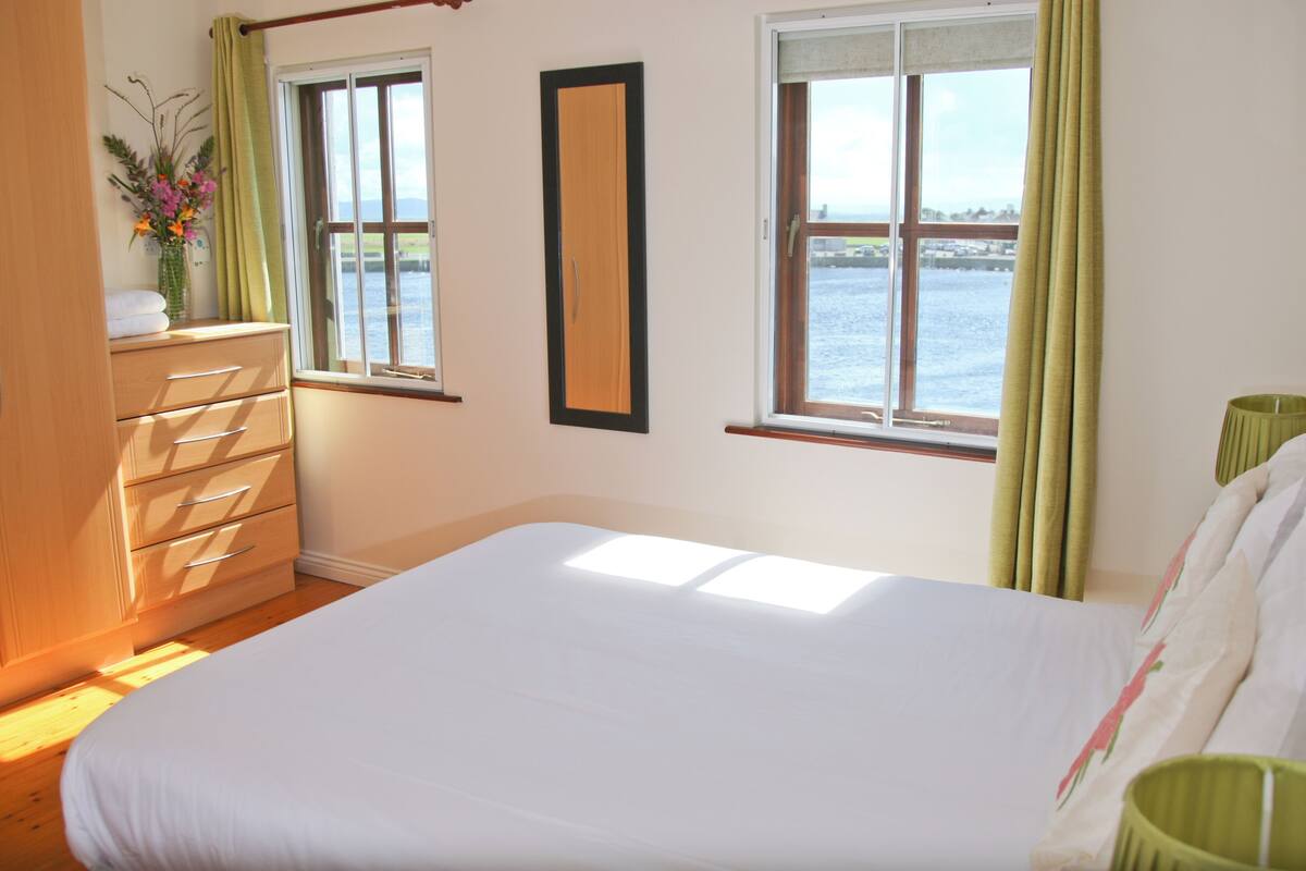 A bedroom with a large bed, a wooden dresser, and two windows with curtains.
