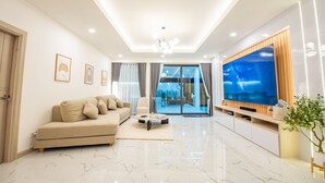 A modern living room with a large flat-screen TV, a beige sofa, a coffee table, and a large window with curtains.