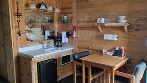 A compact kitchen with a sink, microwave, and coffee maker. There is a wooden table with chairs and a shelf above holding glasses and plates.