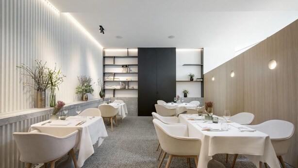 A modern dining area with white tables and chairs, a central black partition, and decorative plants.