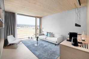 A modern living room with a sofa, a coffee table, a chair, a desk with a coffee machine, and a view of the outdoors.