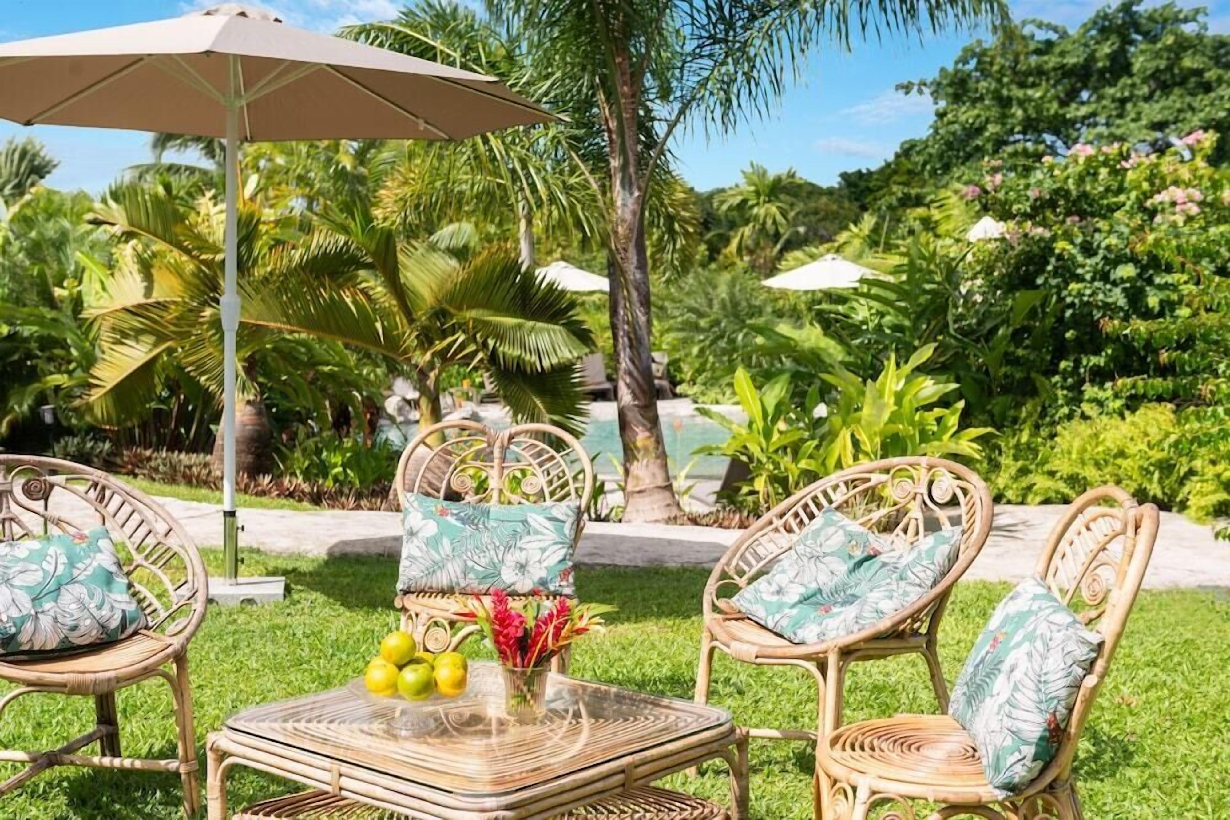 Un espacio al aire libre con ambiente tropical, con sillas de mimbre y una mesa, rodeado de abundante follaje y palmeras.