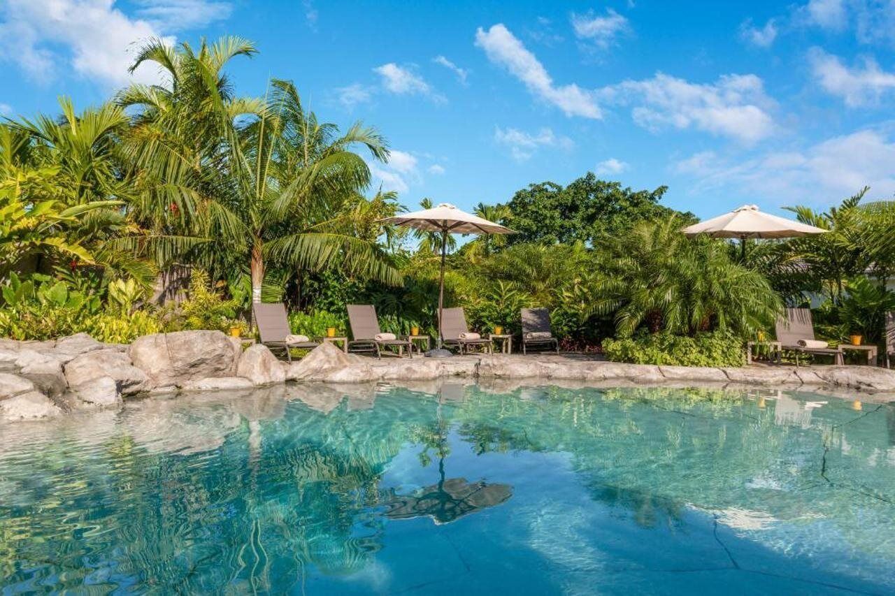 Piscina cristalina con sillas de descanso y sombrillas, rodeada de exuberante vegetación tropical.