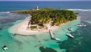 An aerial view of a tropical island with a lighthouse, clear turquoise waters, and several boats.