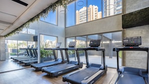 A gym with treadmills, a wall-mounted exercise bike, and large windows offering views of buildings and greenery.