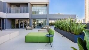 A modern outdoor seating area with a green sofa and a small table, surrounded by planters and a building with large windows and balconies.