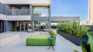 A modern outdoor seating area with a green sofa and a small table, surrounded by planters and a building with large windows and balconies. - Studio w/ Balcony and 290m From the Subway Oln1702 (São Paulo)
