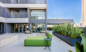 A modern outdoor seating area with a green sofa and a small table, surrounded by planters and a building with large windows and balconies. - Studio w/ Balcony and 290m From the Subway Oln1702 (São Paulo)