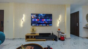 A living room with a flat-screen TV mounted on the wall, a wooden entertainment unit below, and a marble floor. - Elegant 2BR in Lekki Phase 1 • Steady Power, Pool, PS5 , Fast WiFi, Balconies (Lekki)