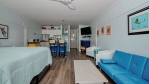 A room with a bed, a blue sofa, a small table, a ceiling fan, a TV, and a kitchen area in the background.
