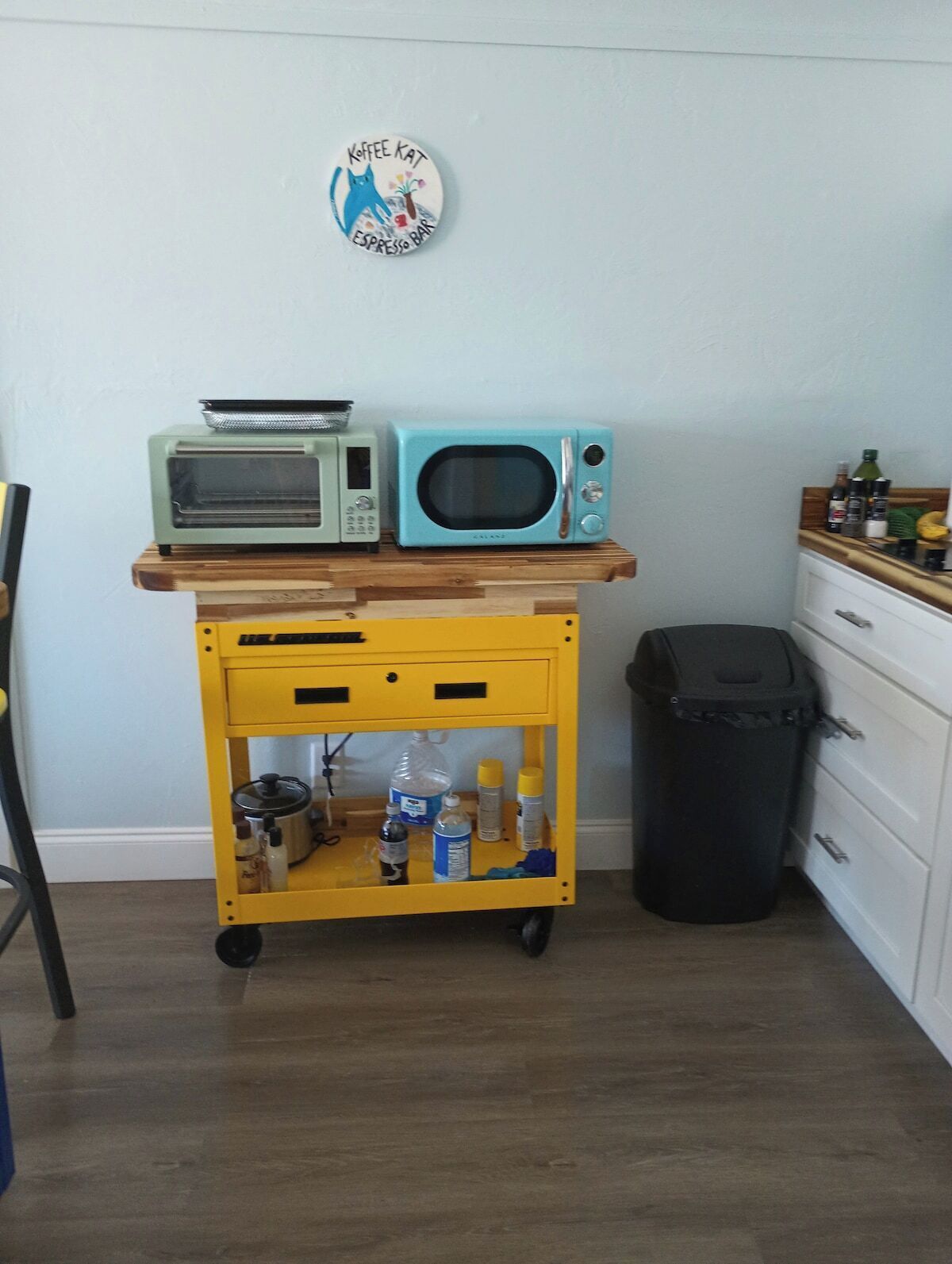 A kitchen with a yellow cart holding a toaster and a microwave, a trash bin, and a wall sign.