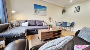 A modern living room with a dark sofa, a wooden coffee table, and a dining area with blue chairs. - Stylish London FLAT near Highgate tube station (London)
