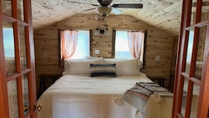 Ein Schlafzimmer einer Holzhütte mit Bett, Vorhängen, Deckenventilator und Fenster.