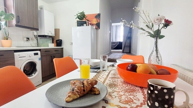 Keittiössä on pyykinpesukone, ruokapöytä oransseilla tuoleilla, lautanen, jossa on croissantteja, kulho täynnä hedelmiä ja maljakko, jossa on kukkia.